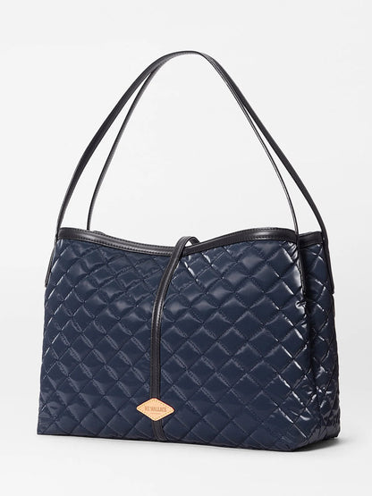 The MZ Wallace Astor East West Tote in Ink Liquid Oxford features a navy blue quilted design, black Italian leather handles, gunmetal hardware, and a tan leather tag, shown against a plain white background.