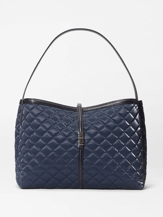The MZ Wallace Astor East West Tote in Ink Liquid Oxford is a navy blue quilted bag with a single black Italian leather shoulder strap and a central vertical black leather accent finished with a metal clasp.