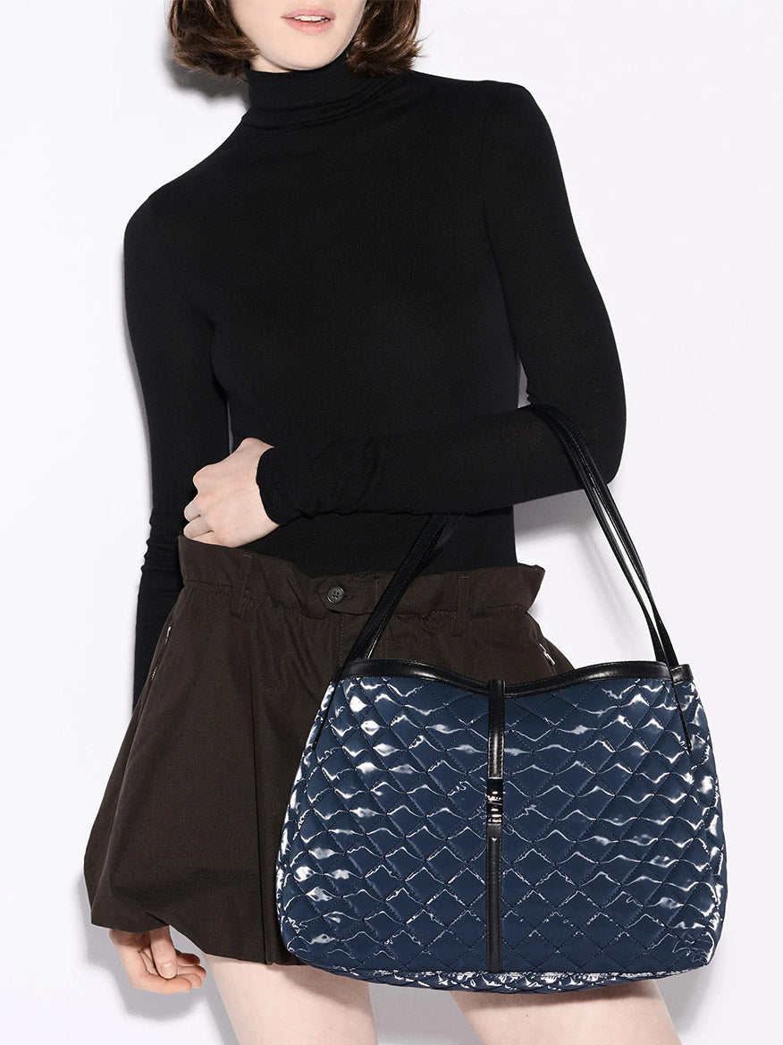 A person in a black turtleneck and brown shorts holds the MZ Wallace Astor East West Tote in Ink Liquid Oxford, featuring quilted blue material, black straps, gunmetal hardware, and zipper details.