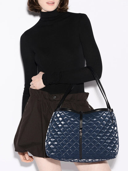 A person in a black turtleneck and brown shorts holds the MZ Wallace Astor East West Tote in Ink Liquid Oxford, featuring quilted blue material, black straps, gunmetal hardware, and zipper details.