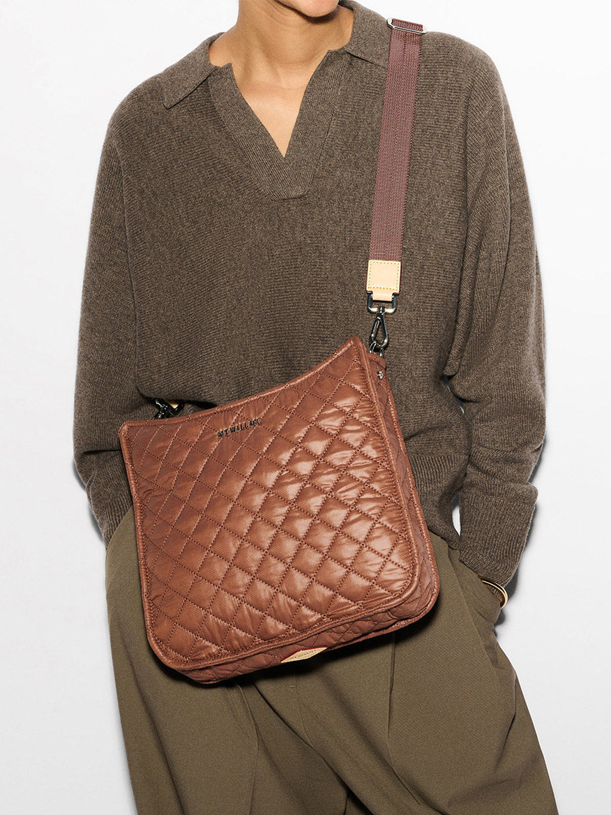 A person wears a brown sweater and pants, gracefully holding the MZ Wallace Metro Box Crossbody in Terracotta Oxford, featuring a quilted design and thick strap.