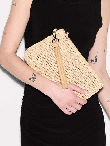 A person in a black sleeveless dress holds the MZ Wallace Crochet Clutch in Crochet/Camel with a leather strap; tattoos visible on their arms highlight this stylish summer accessory.