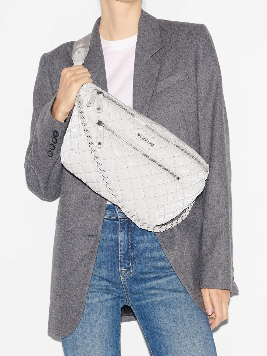 A person in a grey blazer and blue jeans carries a white MZ Wallace Crosby Crossbody Sling Bag in Pebble Liquid with an adjustable nylon crossbody strap.