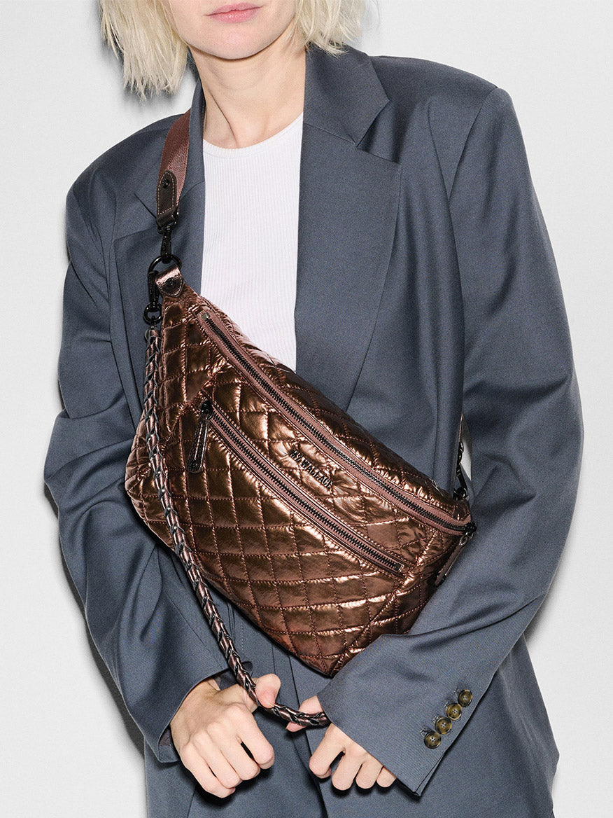 A person wearing a gray blazer and white shirt holds an MZ Wallace Crosby Crossbody Sling Bag in Copper Metallic Lacquer Oxford with Italian leather trim.