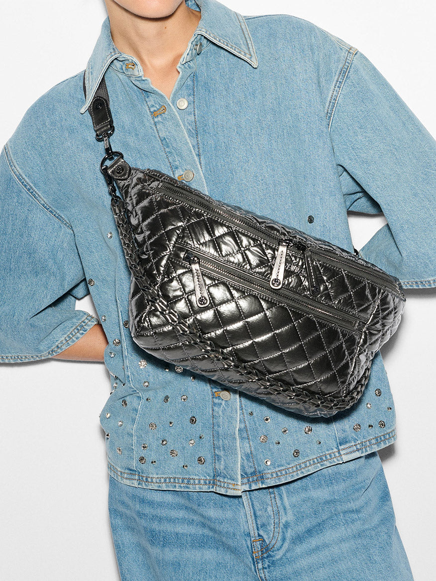 A person sporting a stylish denim jacket and jeans effortlessly carries the MZ Wallace Crosby Crossbody Sling Bag in Pewter Metallic Oxford across their chest, featuring zippers and an adjustable strap for added convenience.