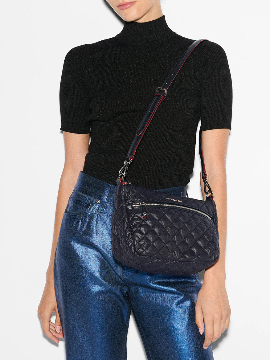 A person wearing a black short-sleeve turtleneck and blue metallic pants, carrying the MZ Wallace Crosby Ella Crossbody in Black Oxford with an elegant adjustable strap.