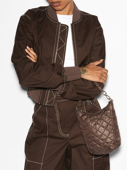 A person is wearing a brown jacket and matching pants with white stitching details, holding an MZ Wallace Crosby Go in Walnut Oxford, a small quilted handbag with an adjustable strap. Their arms are crossed.