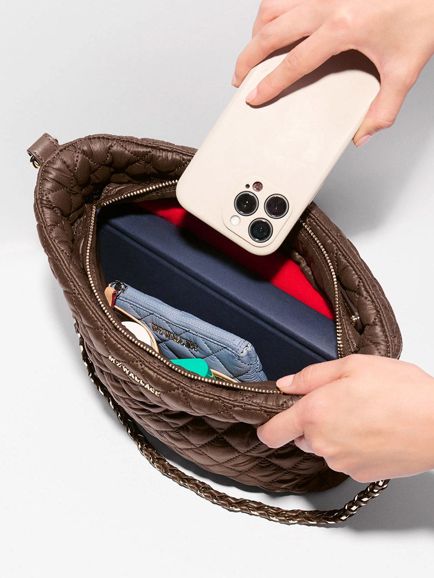 A person puts a smartphone into an open MZ Wallace Crosby Hobo in Walnut Oxford, a brown quilted handbag with Italian leather trim, containing a wallet, pen, and other items.