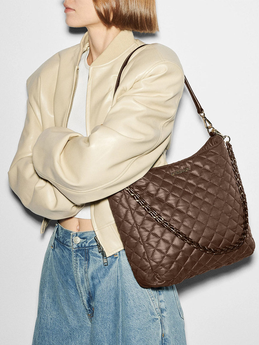 A person in a cream jacket and jeans carries the MZ Wallace Crosby Hobo in Walnut Oxford, a large quilted brown shoulder bag with a chain strap, standing against a plain background.