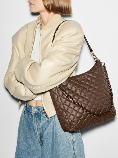 A person in a cream jacket and jeans carries the MZ Wallace Crosby Hobo in Walnut Oxford, a large quilted brown shoulder bag with a chain strap, standing against a plain background.