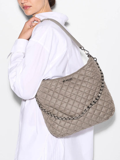 A woman in a white shirt carries the spacious MZ Wallace Crosby Hobo in Morning Dove Oxford, a quilted beige shoulder bag made from recycled nylon with Italian leather accents and a braided strap.