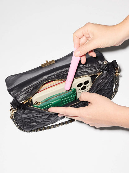 Hands placing a pink tube into an MZ Wallace Crosby Lock Baguette in Black Oxford containing a smartphone and a green wallet.