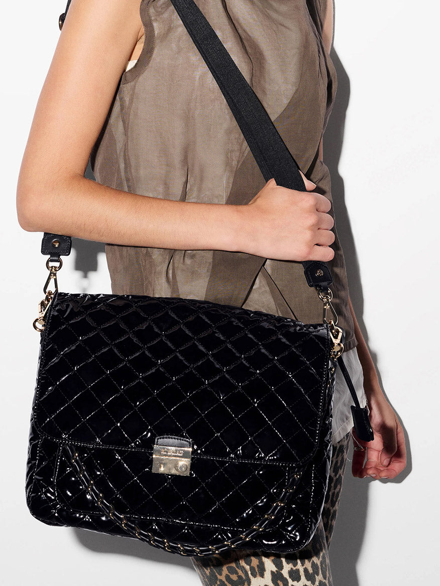 A person in a sheer brown sleeveless top and leopard print pants holds the MZ Wallace Crosby Lock Large Crossbody in Black Lacquer Oxford.