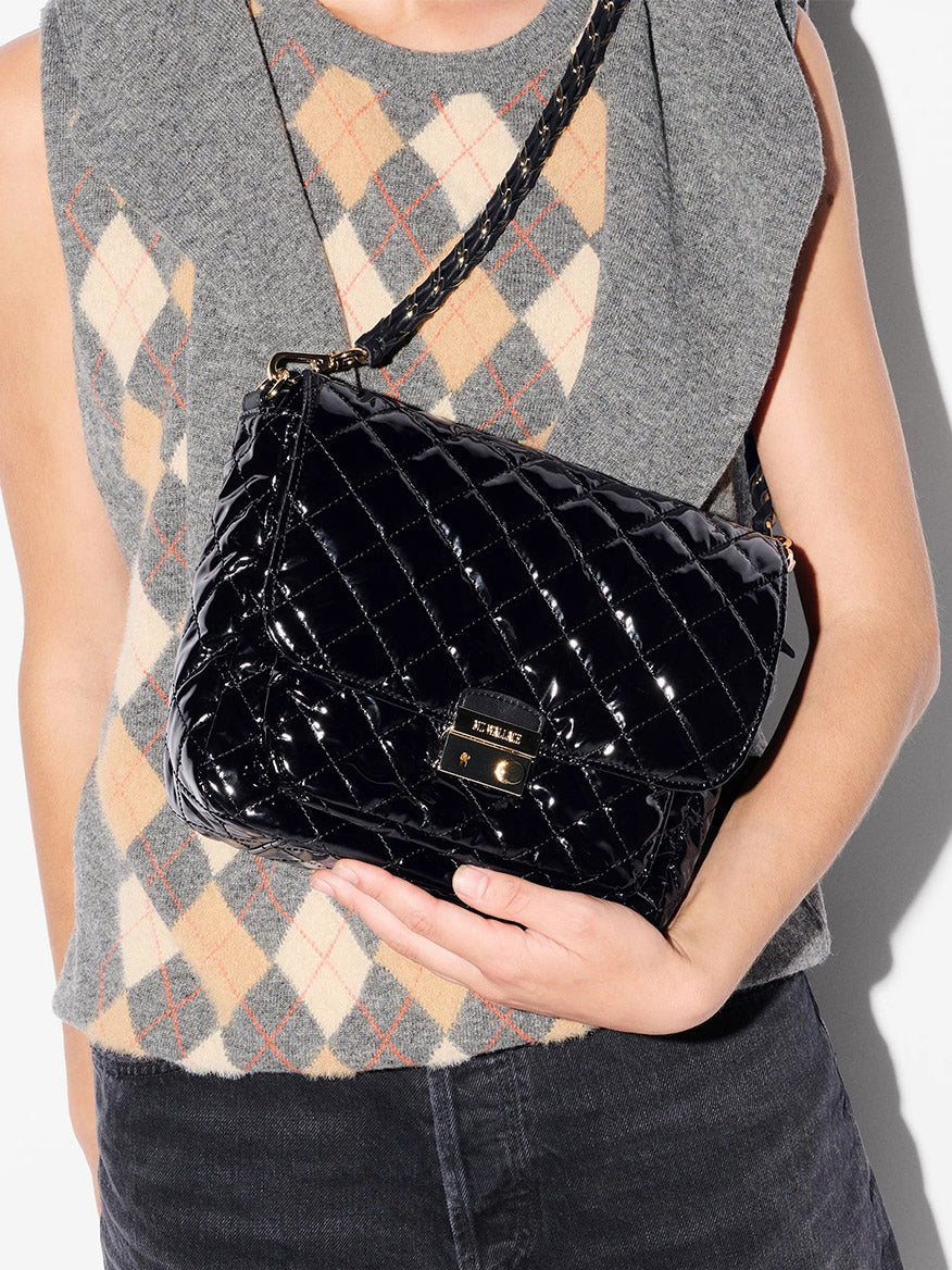 A person in a gray argyle sweater holds the MZ Wallace Crosby Lock Medium Crossbody in Black Lacquer Oxford—a sleek, quilted designer bag with a gold clasp and shoulder strap.