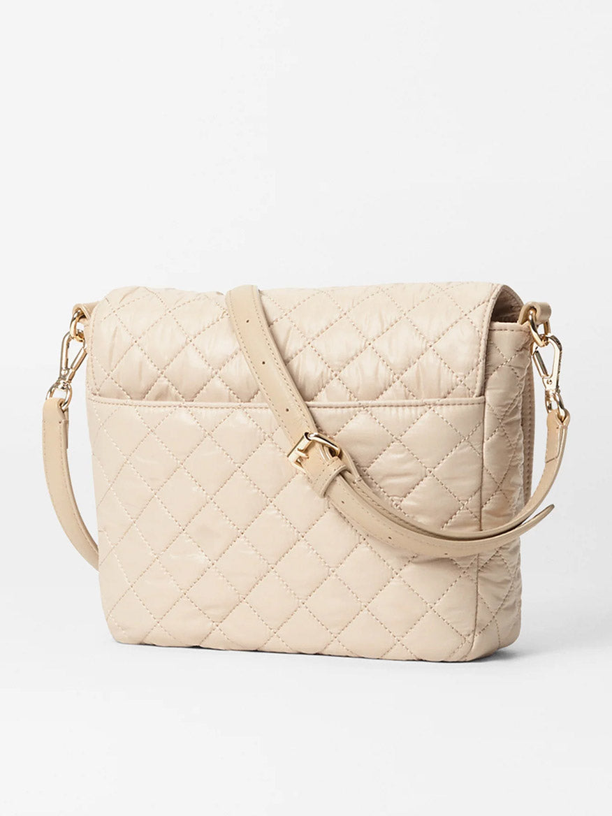 The MZ Wallace Crosby Lock Medium Crossbody in Buff Oxford is a beige nylon quilted shoulder bag featuring an adjustable strap, gold-tone hardware, and a fold-over flap design, embodying elegance and serving as a true designer masterpiece against a white background.