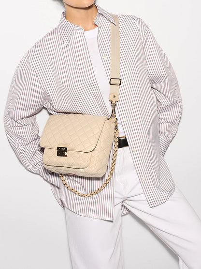 A person is dressed in a white and blue striped shirt with a white tee underneath, paired with white trousers. They have the MZ Wallace Crosby Lock Medium Crossbody in Buff Oxford, featuring a nylon quilted finish and chain strap, over their shoulder.