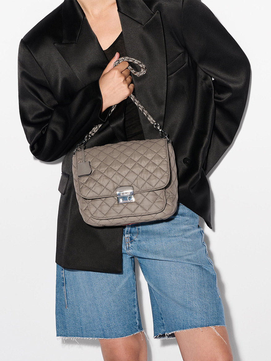 Person wearing a black blazer and denim shorts, holding the MZ Wallace Crosby Lock Medium Crossbody in Morning Dove Oxford, a stylish quilted nylon designer bag with chain strap and metal clasp.
