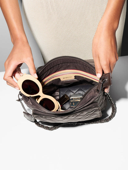 Someone places cream-colored sunglasses into an open MZ Wallace Crosby Lock Medium Crossbody in Espresso Oxford, which also holds a lipstick and a small black item, all set on a white surface.