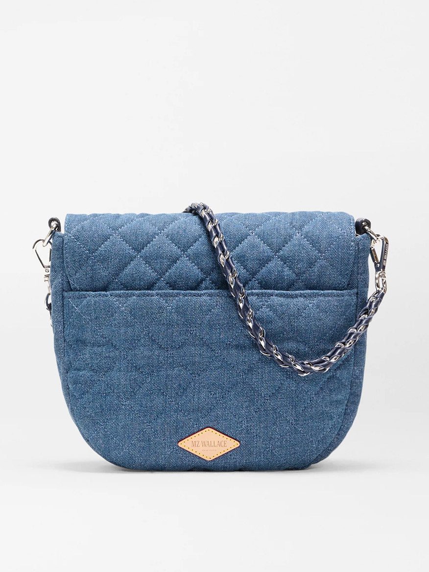 The MZ Wallace Crosby Lock Saddle in True Blue Denim features a blue quilted design, Italian leather trim, chain strap, and a small leather logo patch on the front, shown against a plain white background.