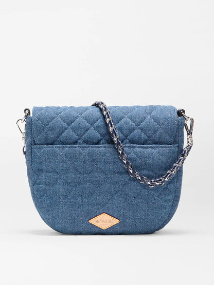The MZ Wallace Crosby Lock Saddle in True Blue Denim features a blue quilted design, Italian leather trim, chain strap, and a small leather logo patch on the front, shown against a plain white background.