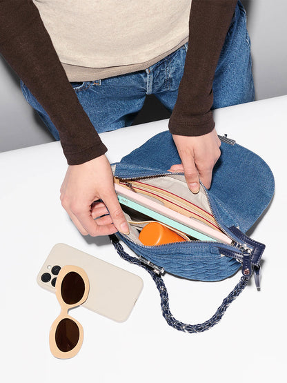 A person puts a notebook and orange container into the MZ Wallace Crosby Lock Saddle in True Blue Denim, set on a white surface next to sunglasses and a smartphone.