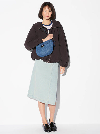 A woman stands against a white background wearing a dark jacket, light denim skirt, black loafers, and carrying the MZ Wallace Crosby Lock Saddle in True Blue Denim with Italian leather trim.