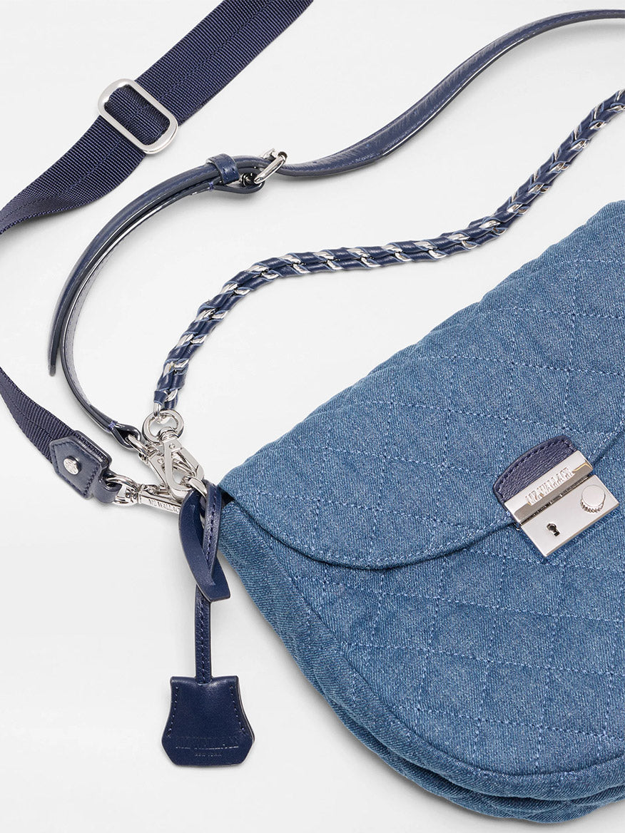 The MZ Wallace Crosby Lock Saddle in True Blue Denim features a blue quilted design, silver buckle, Italian leather trim, braided handle, leather tag, and adjustable strap displayed on a white surface.