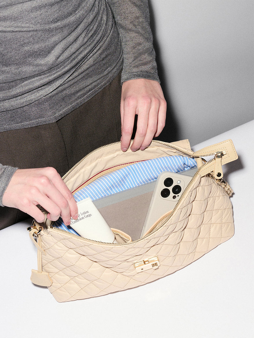 A person puts a white smartphone and a small book into an MZ Wallace Crosby Lock Shoulder in Buff Oxford, featuring blue-and-white striped lining and interchangeable straps.