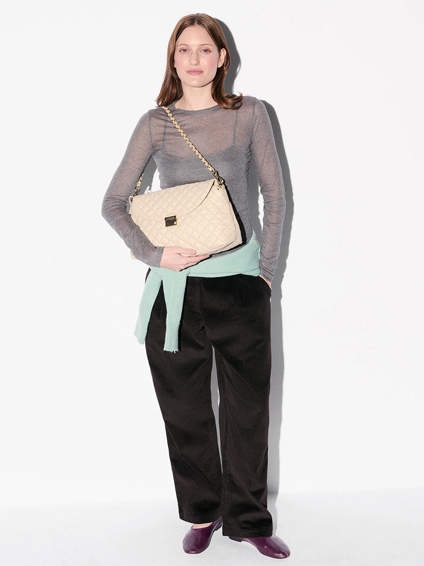 A woman stands against a white background wearing a sheer gray top, black pants, and purple shoes. She holds the MZ Wallace Crosby Lock Shoulder in Buff Oxford with interchangeable straps and has a light sweater tied at her waist.