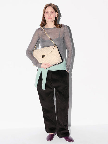 A woman stands against a white background wearing a sheer gray top, black pants, and purple shoes. She holds the MZ Wallace Crosby Lock Shoulder in Buff Oxford with interchangeable straps and has a light sweater tied at her waist.