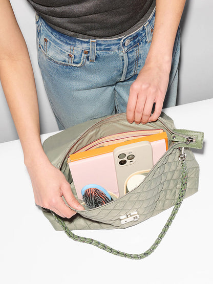 A person opens the MZ Wallace Crosby Lock Shoulder in Agave Green Oxford, revealing a phone, notebook, yellow folder, round mirror, and hairbrush inside.