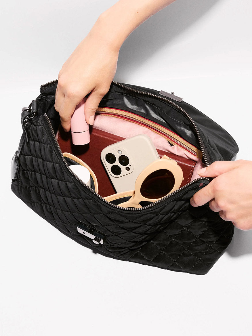 Hands opening a black MZ Wallace Crosby Lock Shoulder in Black Velvet Finish to reveal a smartphone, sunglasses, and personal items inside.