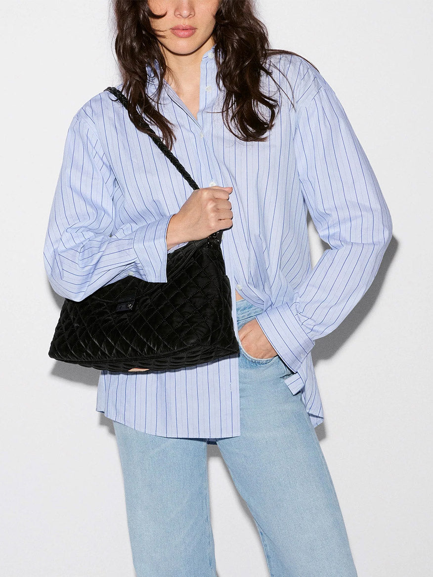 A person wearing a blue striped button-down shirt and light wash jeans carries a black velvet MZ Wallace Crosby Lock Shoulder bag with a braided strap.