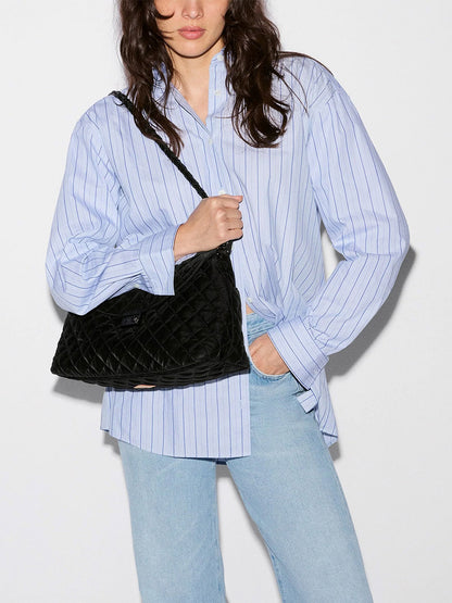A person wearing a blue striped button-down shirt and light wash jeans carries a black velvet MZ Wallace Crosby Lock Shoulder bag with a braided strap.