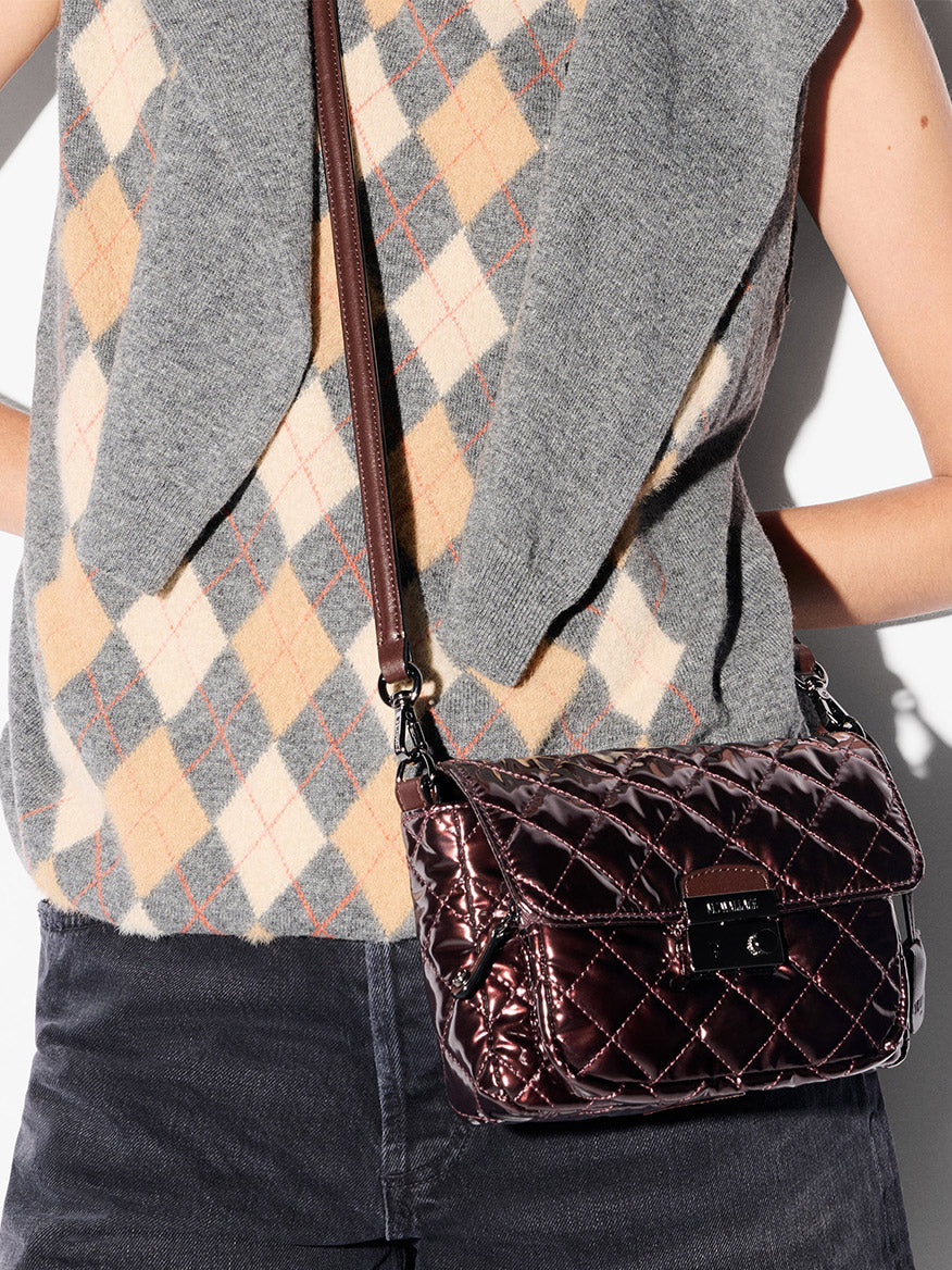 A person in a grey argyle sweater vest and dark pants carries an MZ Wallace Crosby Lock Small Crossbody in Bordeaux Lacquer Oxford.
