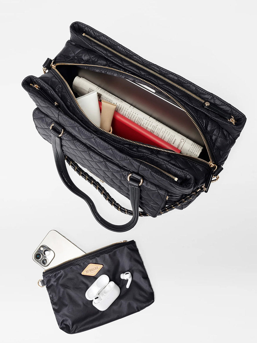Top view of the MZ Wallace Crosby Lock Tote in Black Oxford with its main compartment partially open, revealing books and papers inside. Below it is a small black pouch containing an iPhone, AirPods, and a lipstick. This stylish nylon work tote is perfect for organizing all your essentials on the go.