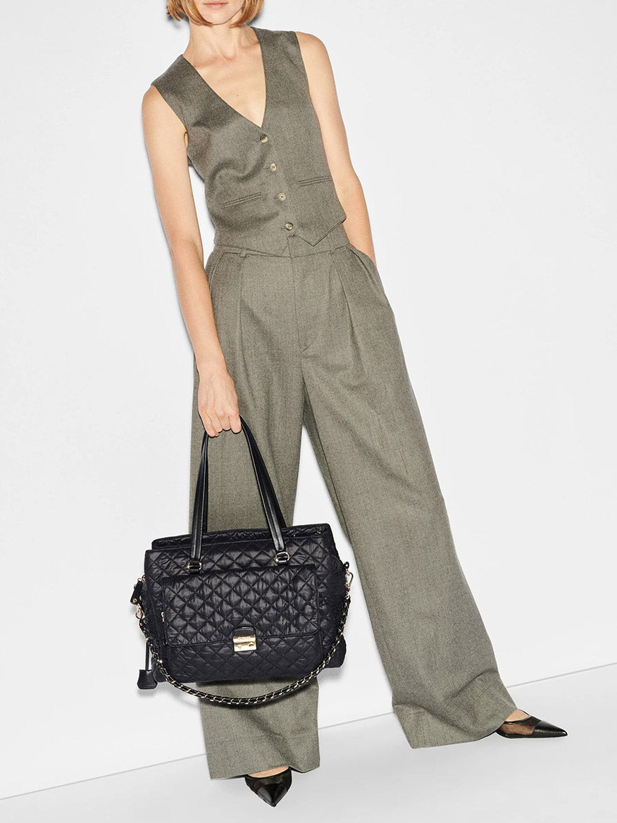 A person wearing a sleeveless, buttoned grey jumpsuit holds a designer work bag—a quilted MZ Wallace Crosby Lock Tote in Black Oxford with chain handles—set against a plain white background.