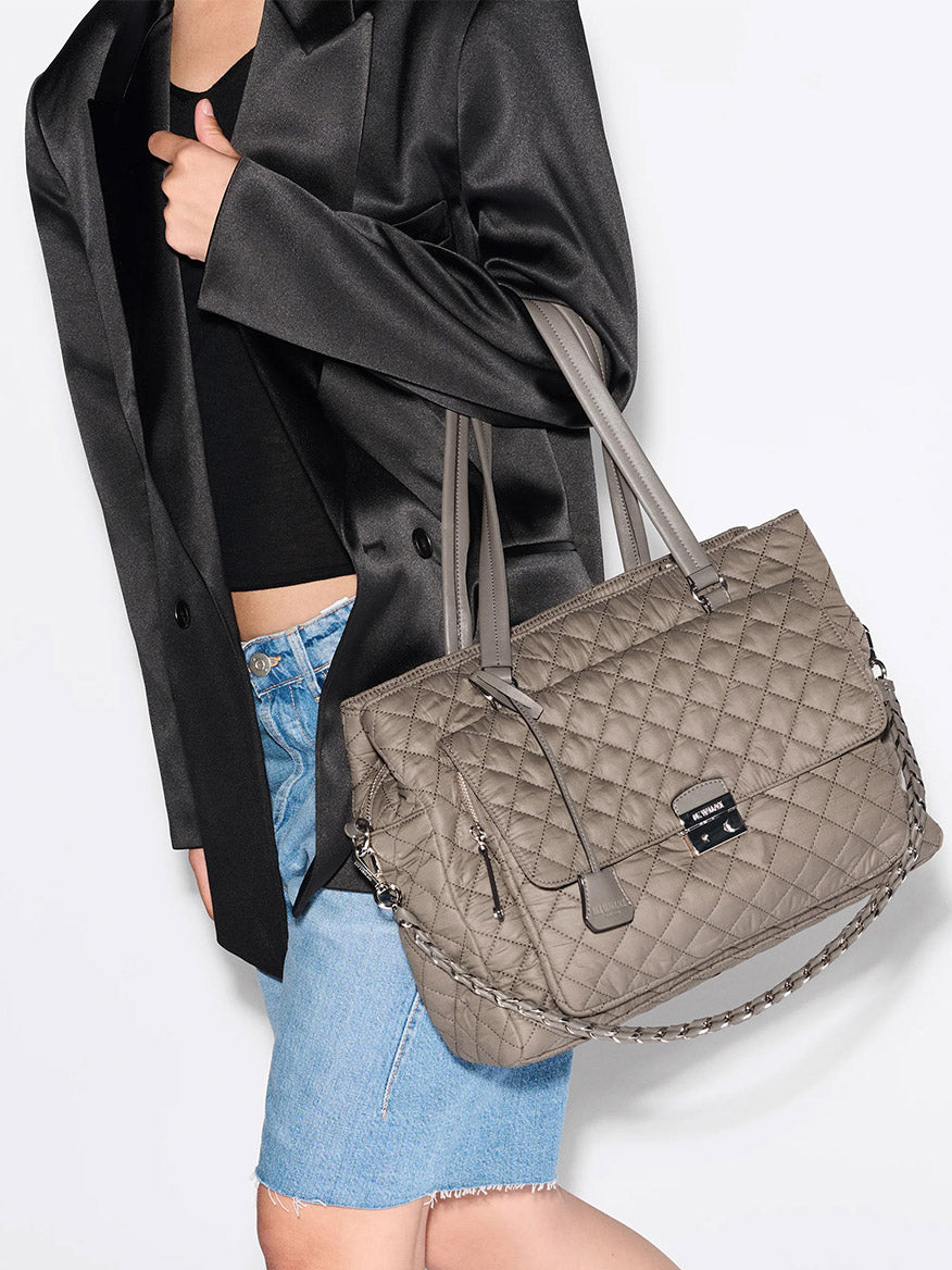 A person wearing a black blazer, black top, and denim skirt carries the MZ Wallace Crosby Lock Tote in Morning Dove Oxford—a quilted taupe tote with double handles and a chain strap.