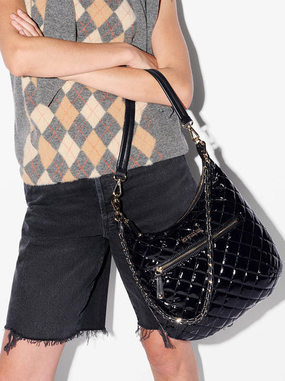 Person wearing a grey argyle sweater vest and black denim shorts with frayed hems, holding the MZ Wallace Crosby Sienna in Black Lacquer Oxford—a large quilted shoulder bag with gold hardware and Italian leather trim.