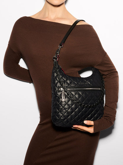 A person in a brown dress holds the MZ Wallace Crosby Sienna in Black Oxford, a black quilted handbag crafted from Italian leather, with a zipper pocket and shoulder strap.