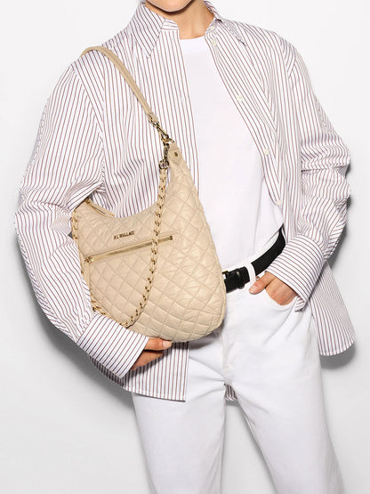 Elegantly dressed in a striped shirt and white pants, the person carries the MZ Wallace Crosby Sienna in Buff Oxford— a quilted beige handbag with a chic chain strap featuring gleaming gold hardware.