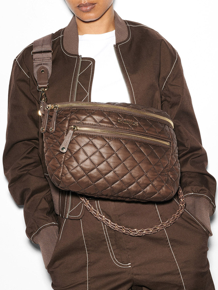 Person wearing a brown bomber jacket and an MZ Wallace Crosby Crossbody Sling Bag in Walnut Oxford with gold zippers.