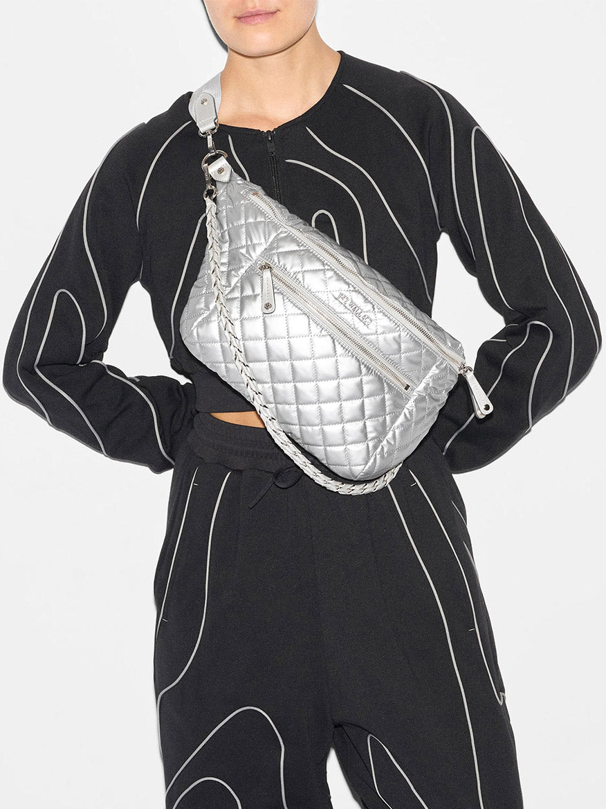 A person wearing a black outfit with white outlines is holding the MZ Wallace Crosby Crossbody Sling Bag in Matte Silver Oxford, which features a silver quilted design and a braided strap across their chest. This versatile, hands-free option offers multiple ways to wear it, making it perfect for any occasion.