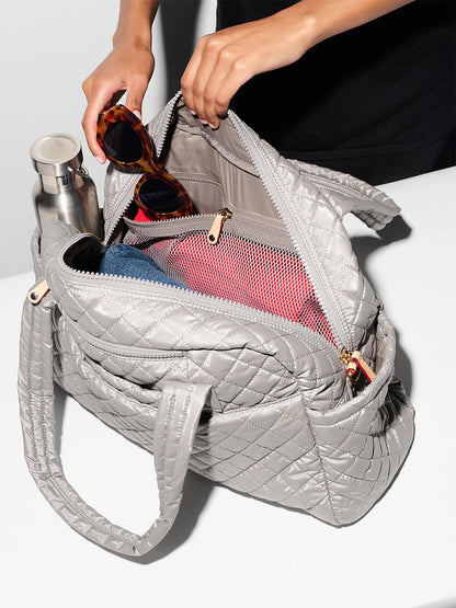 A person puts sunglasses into an open MZ Wallace Jimmy Deluxe in Sable Pearl Metallic, a gray quilted bag with Italian leather trim, which already contains a water bottle and rolled clothes.