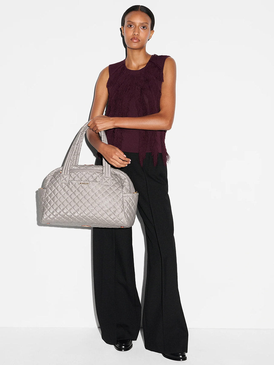 A woman stands against a white background wearing a sleeveless burgundy top, black wide-leg pants, black shoes, and holding the MZ Wallace Jimmy Deluxe in Sable Pearl Metallic with Italian leather trim.