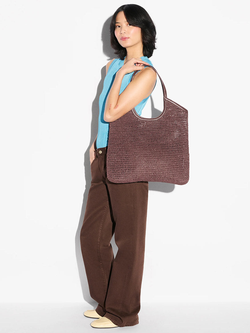 A woman stands against a white background, wearing a blue sleeveless top, brown pants, cream shoes, and carrying the MZ Wallace Large Crochet Georgica Tote in Brown Crochet on her shoulder.