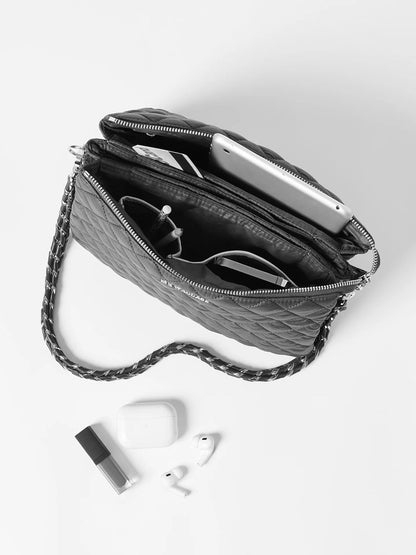 A black quilted MZ Wallace Large Crosby Pippa in Light Gold Pearl Metallic Oxford with an adjustable chain strap is open, revealing compartments holding an iPad, glasses, pens, and headphones. On a white surface next to it are lip balm, wireless earphones, and a charging case. The bag's Italian leather trim adds a touch of elegance to the versatile design.