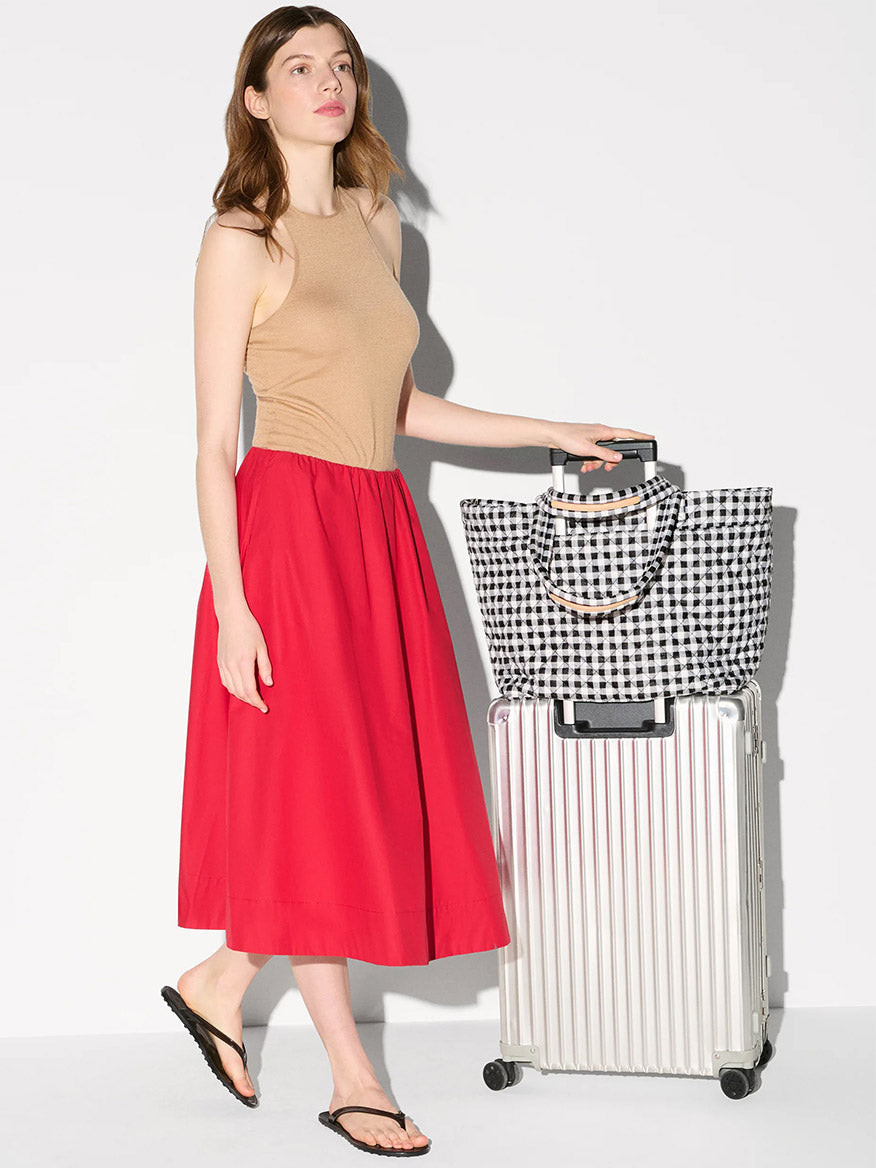 A woman in a beige sleeveless top, red skirt, and black sandals stands beside a silver suitcase topped with the MZ Wallace Large Metro Tote Deluxe in Black/White Gingham Oxford.