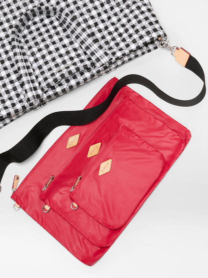 The MZ Wallace Large Metro Tote Deluxe in Black/White Gingham Oxford is partially shown beside a vivid red crossbody with tan leather accents and a black adjustable strap on a white surface—ideal for travel.