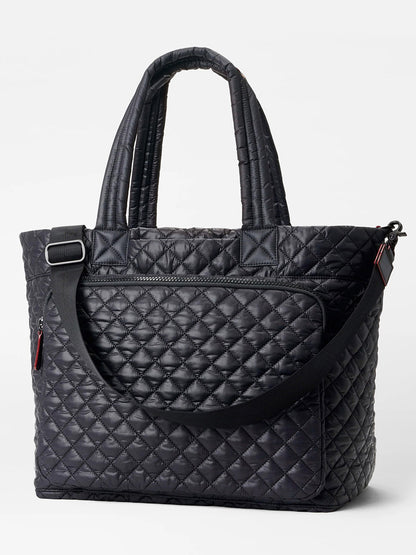 The MZ Wallace Large Metro Travel Tote in Black Oxford features a quilted design, dual handles, Italian leather trim, a front zip pocket, and an adjustable shoulder strap, displayed against a plain light background.